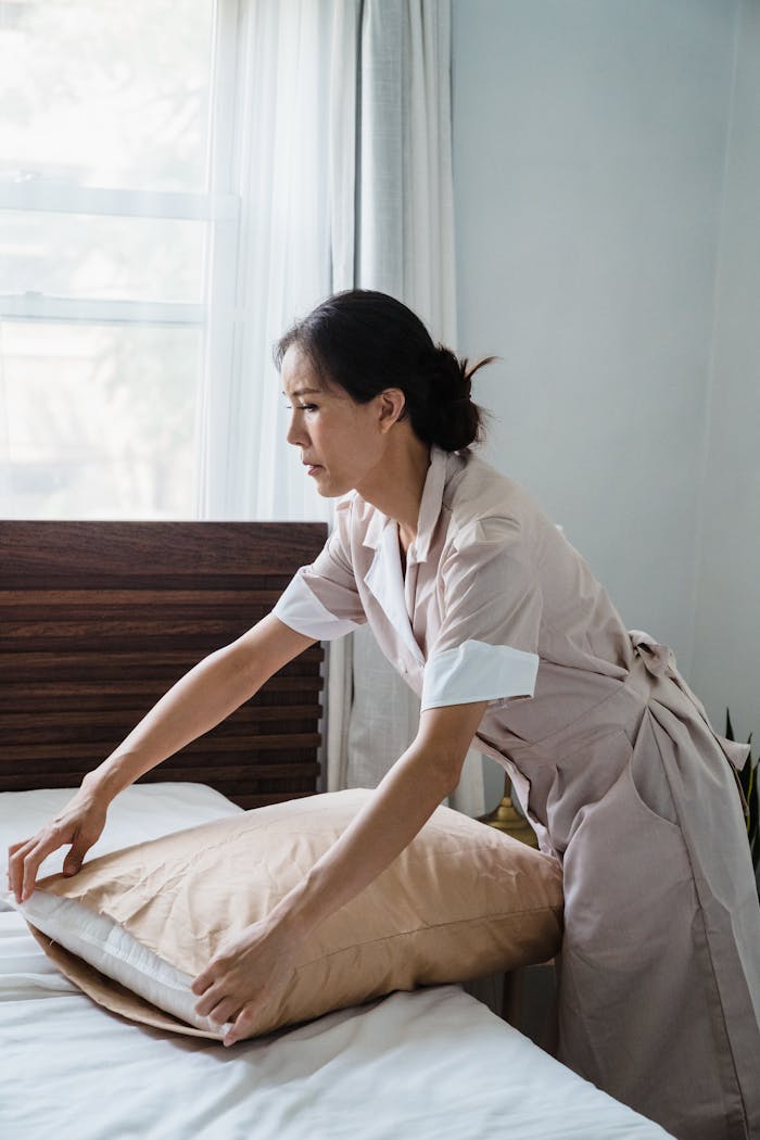 hero-img-02 Asian woman in a tan uniform arranging a pillow in a bright, sunlit bedroom.