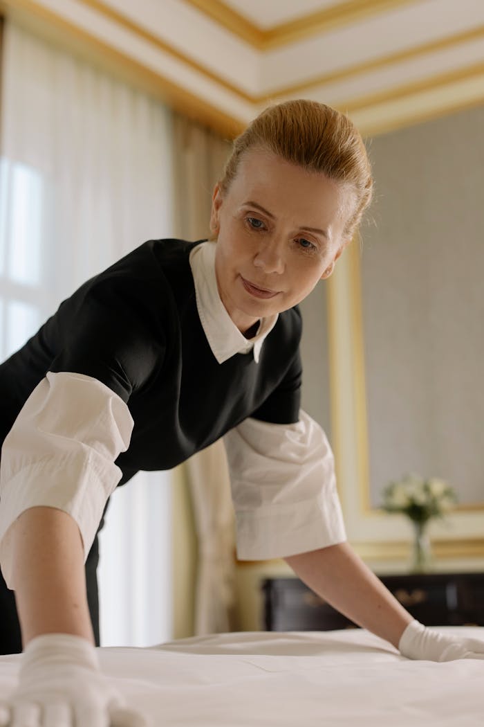 about-01 A housekeeper in uniform skillfully arranges bed linen in a luxurious hotel room.